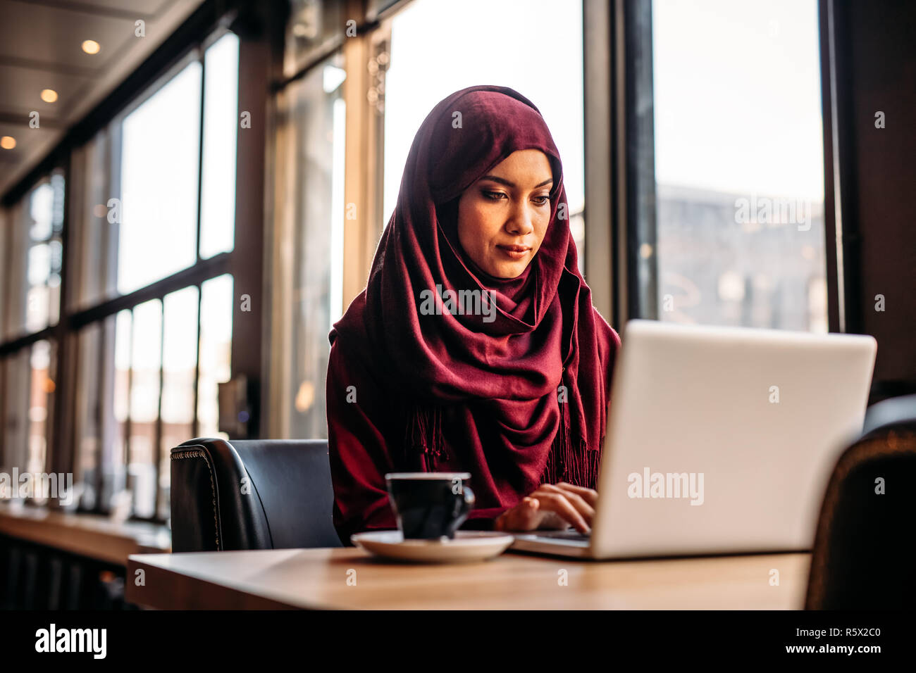 Bella donna islamica seduta al tavolo del caffè e lavorava con un computer portatile con un caffè a lato. donna d'affari in hijab che lavora da un bar. Foto Stock