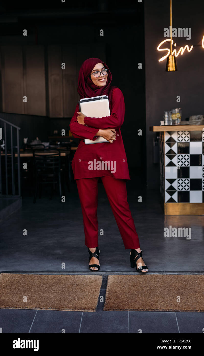 Donna islamica indossano hijab tenendo il computer portatile e guardando lontano. Per tutta la lunghezza della donna musulmana in piedi presso la caffetteria porta con laptop. Foto Stock
