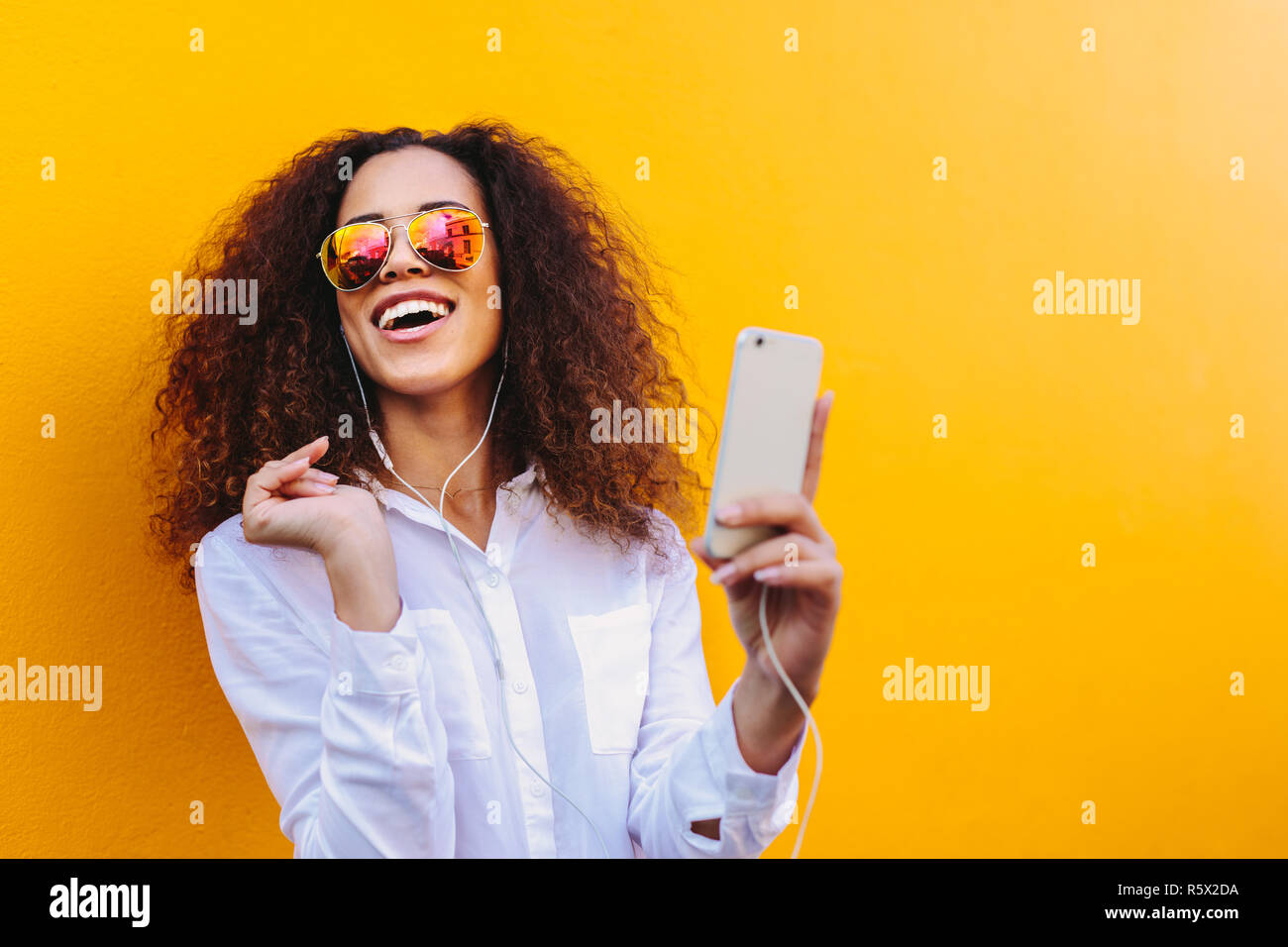 Felice giovane donna africana ascoltando musica in cuffia utilizzando un telefono cellulare. La ragazza con i capelli ricci indossando occhiali da sole contro la parete gialla godendo l Foto Stock