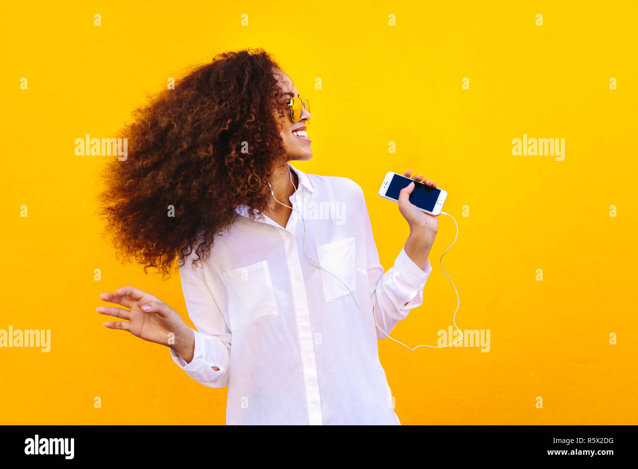 Donna con capelli ricci che indossa una camicia bianca ascoltando la musica dal suo telefono cellulare. Ragazza africana utilizzando auricolari per ascoltare musica contro la parete gialla. Foto Stock