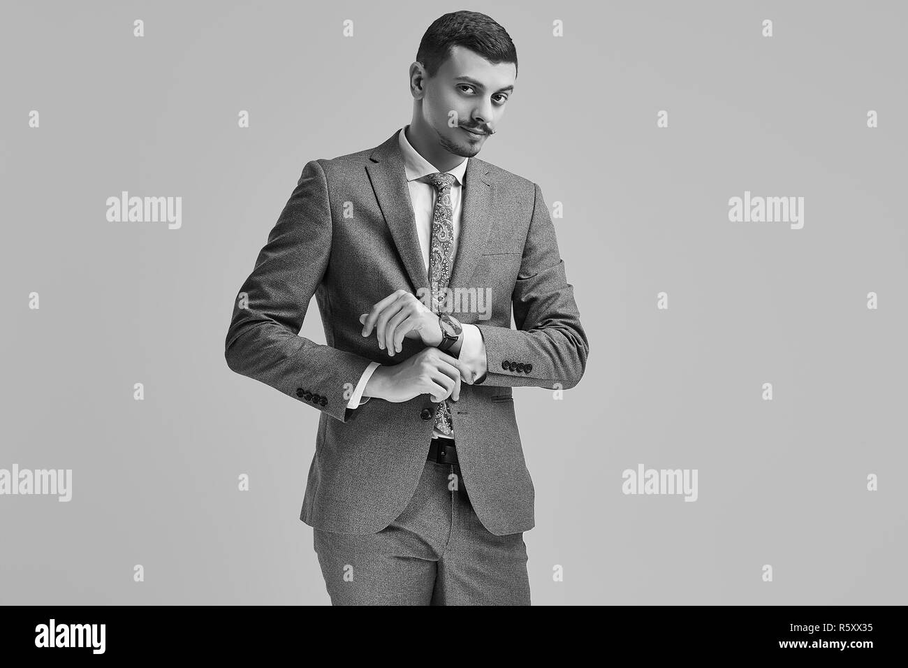 Ritratto di bel giovane fiducioso arabo imprenditore con baffi di fantasia in moda grigio tuta completa la regolazione del suo manicotto su sfondo per studio Foto Stock