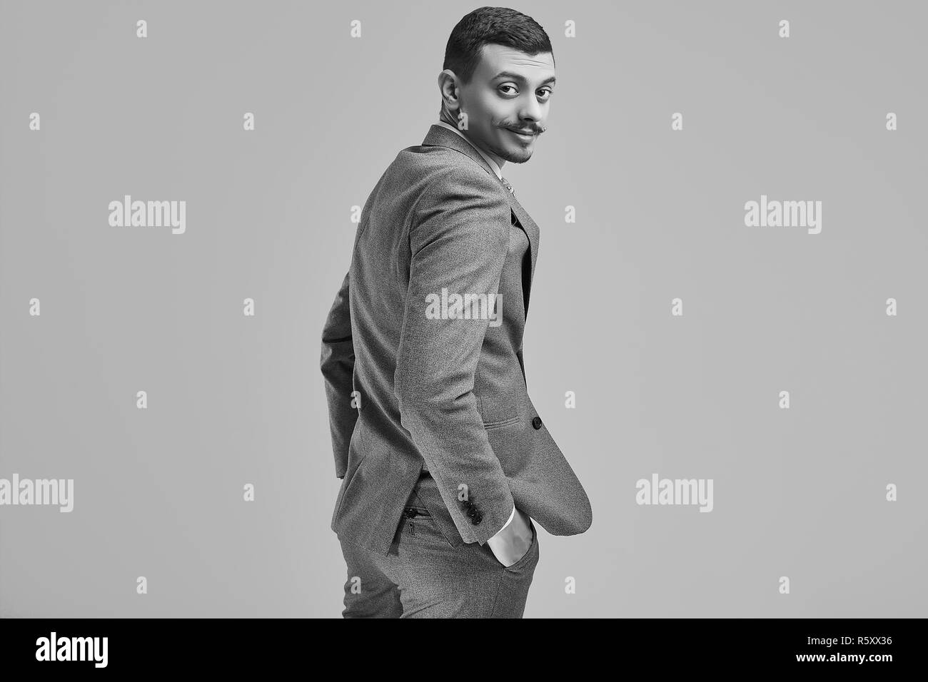 Ritratto di bel giovane fiducioso arabo imprenditore con baffi di fantasia in moda grigio tuta completa appare sopra la sua spalla su sfondo per studio Foto Stock