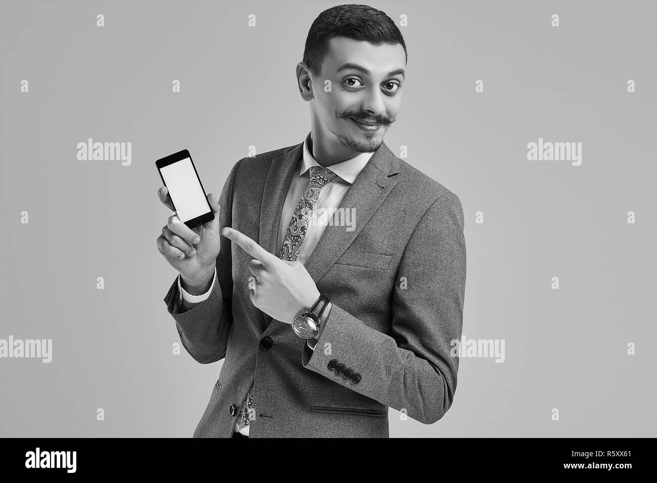 Ritratto di bel giovane fiducioso arabo imprenditore con baffi di fantasia in moda grigio tuta completa di punti al telefono su sfondo per studio Foto Stock