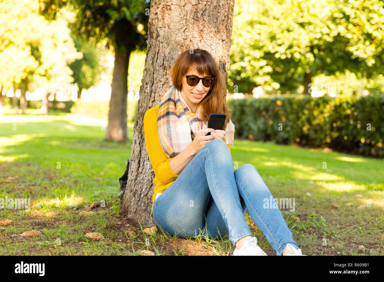 Felice giovane donna seduta accanto ad albero e la digitazione di messaggi sul telefono cellulare. Foto Stock