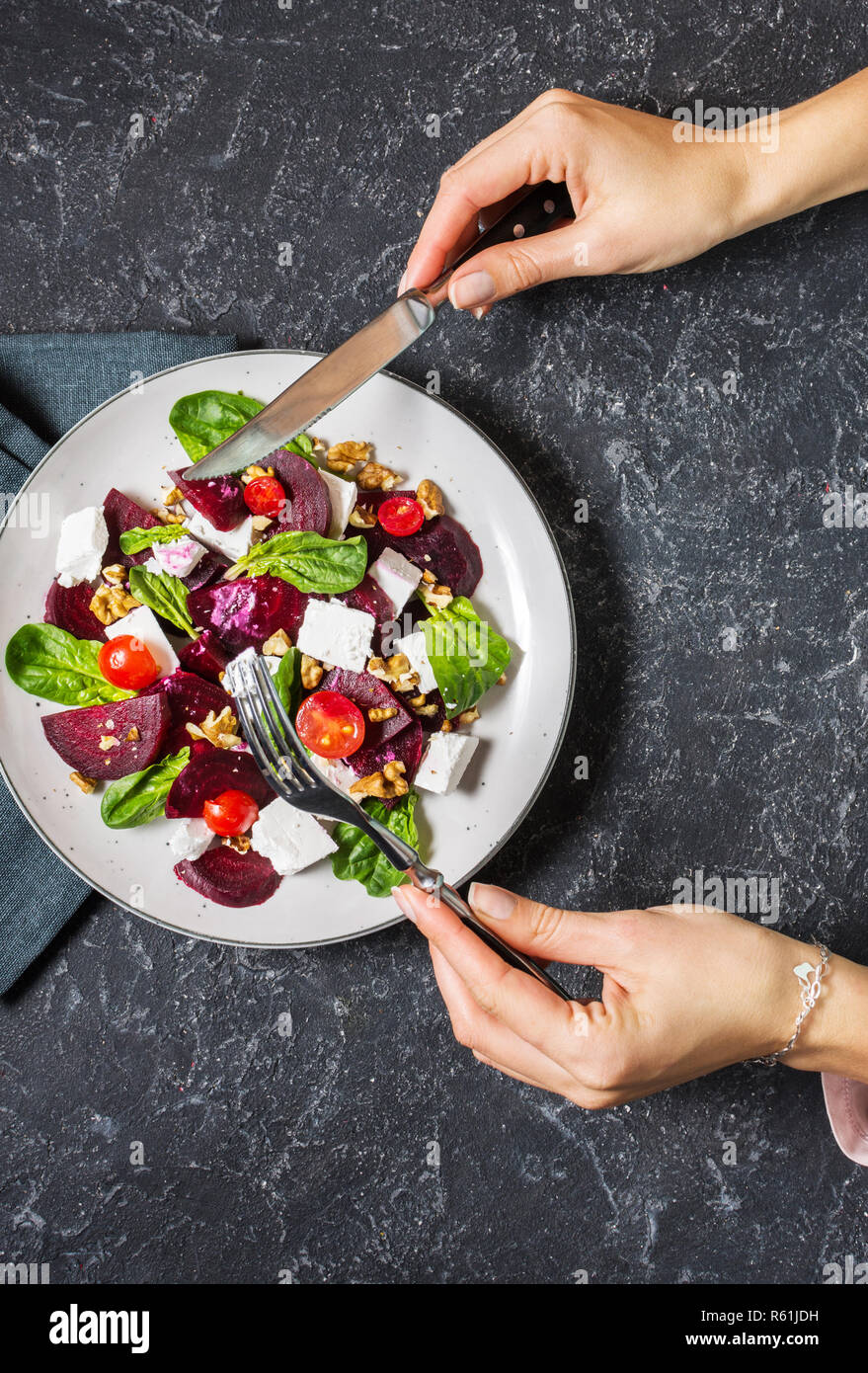 Il piatto con una insalata di feta, barbabietole, pomodorini e noci su un sfondo di pietra. Mani femminili tenere forchetta e coltello. Vista superiore Foto Stock