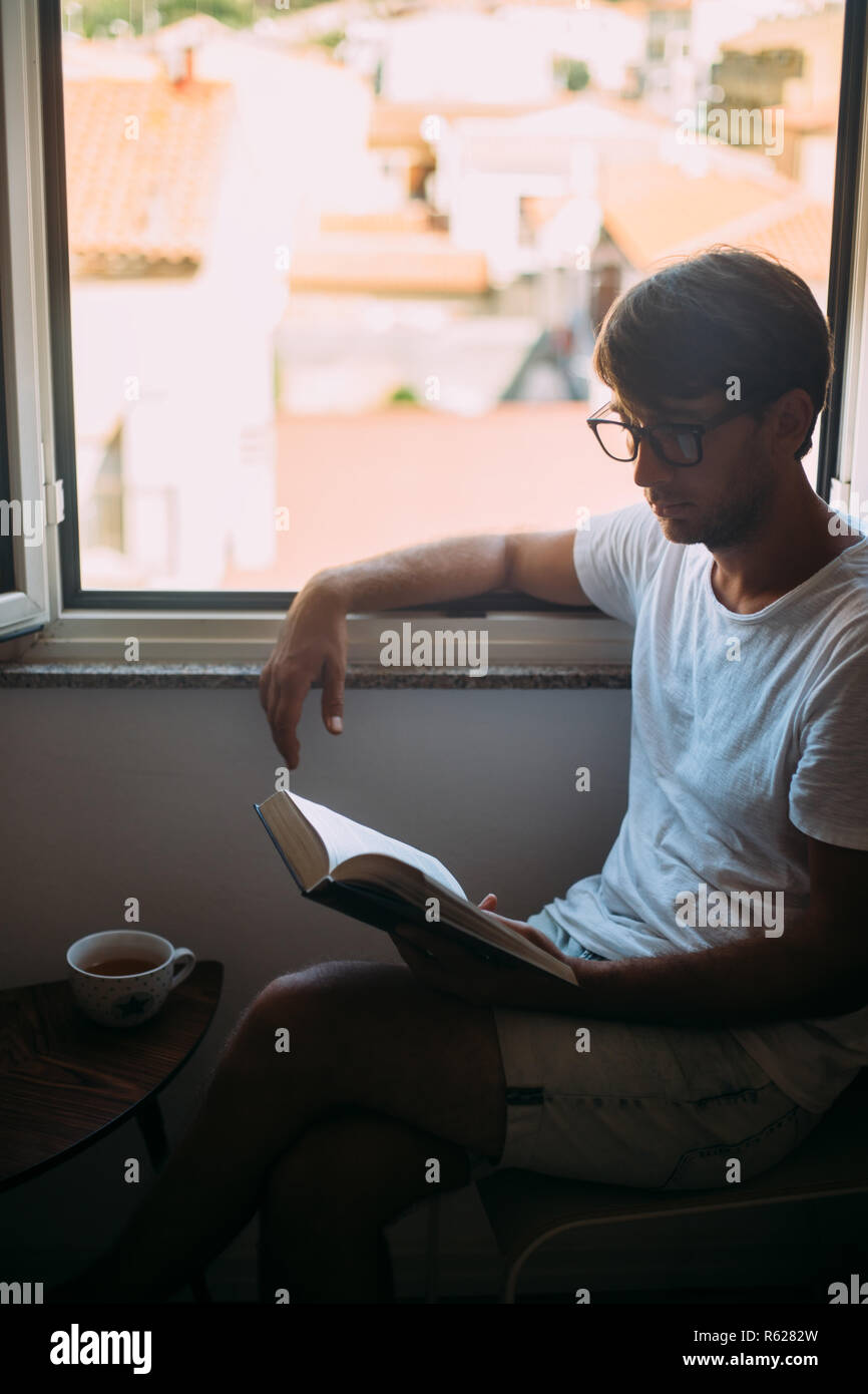 Giovane uomo bello in occhiali lettura prenota vicino alla finestra. weekend concept Foto Stock