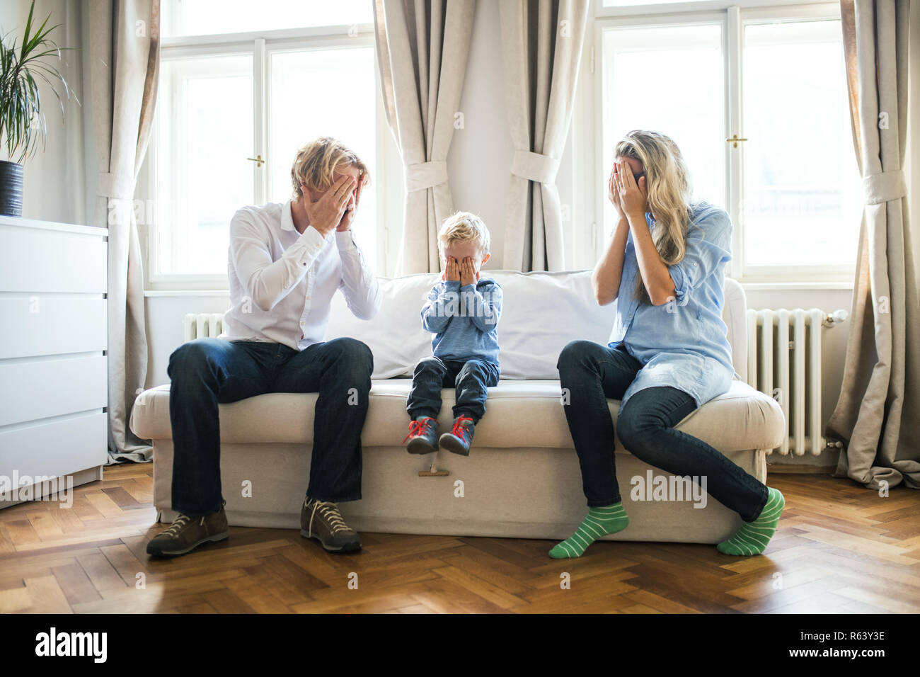 I giovani genitori e bimbo figlio seduto su un divano interno in una camera da letto, che copre gli occhi. Foto Stock