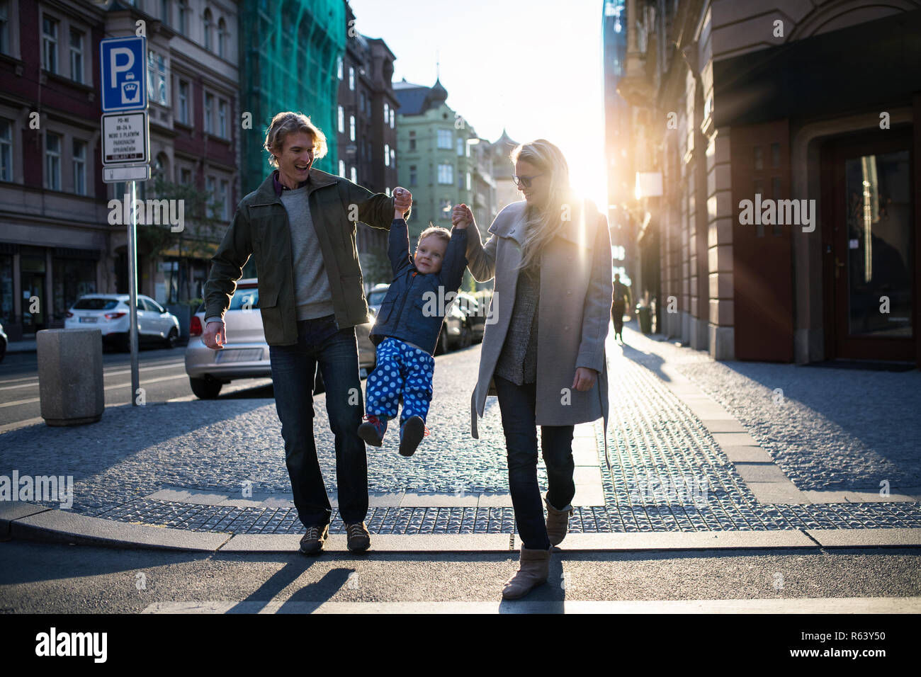 Un piccolo bimbo ragazzo con i genitori attraversare una strada all'aperto in città al tramonto. Foto Stock