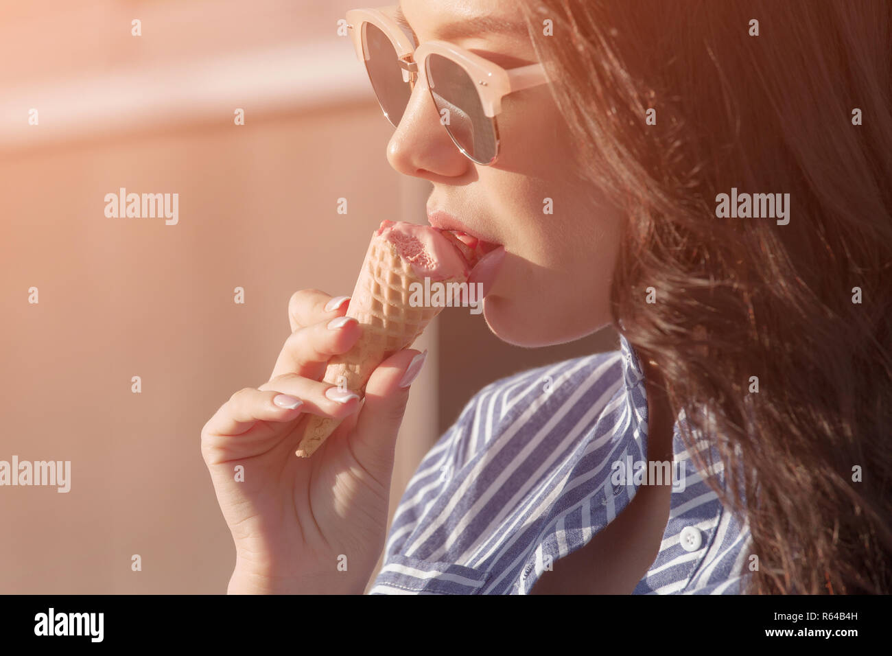 Giovane e bella bruna donna rosa di mangiare il gelato in una cialda avvisatore acustico. Indossa occhiali con occhiali scuri e una camicia in un bianco con striscia blu Foto Stock