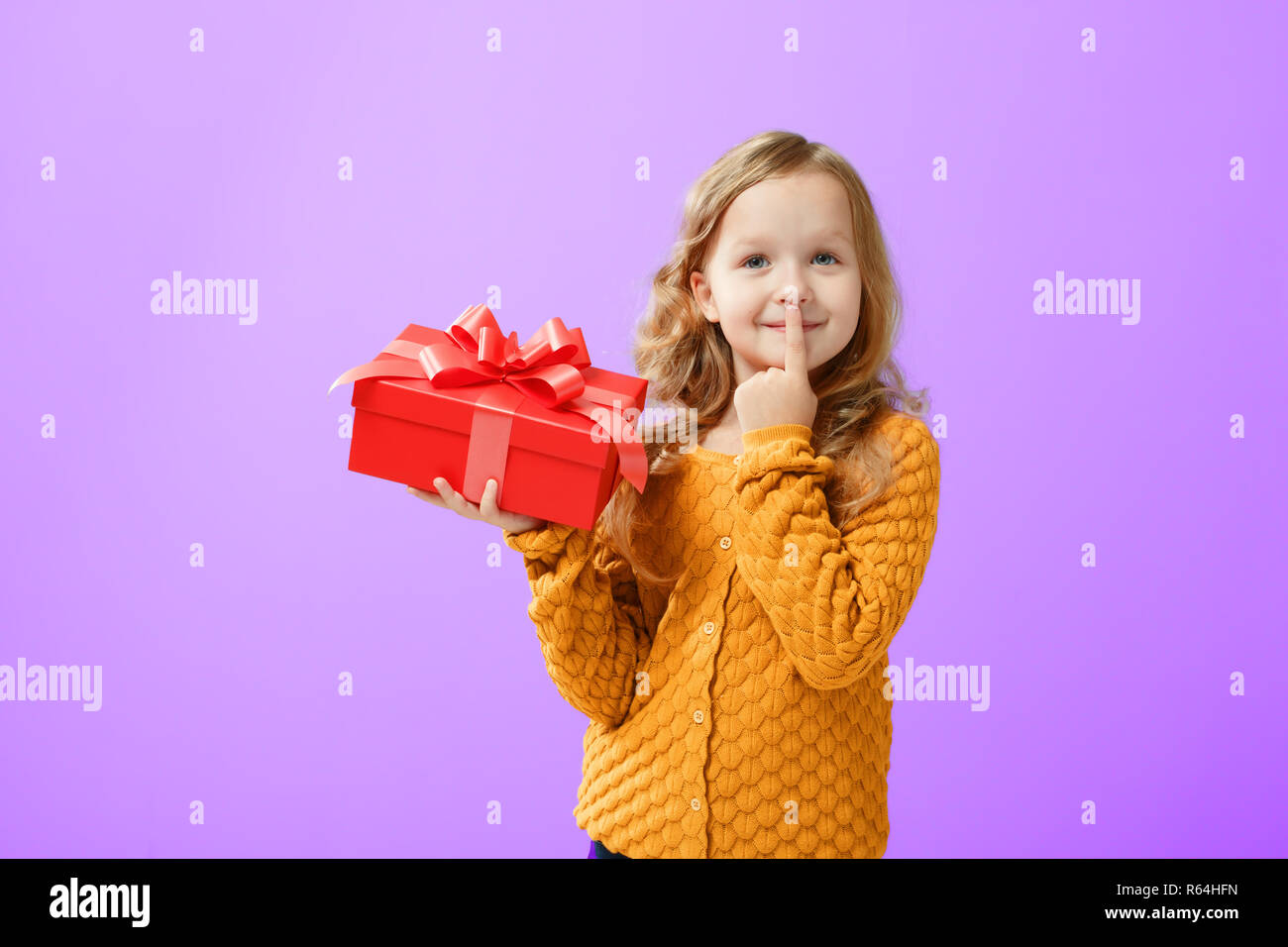 Ritratto di una ragazzina in un maglione caldo su un protone sfondo viola. Il Bambino guarda dreamily fino e contiene una casella rossa con un regalo. Il concetto di Foto Stock