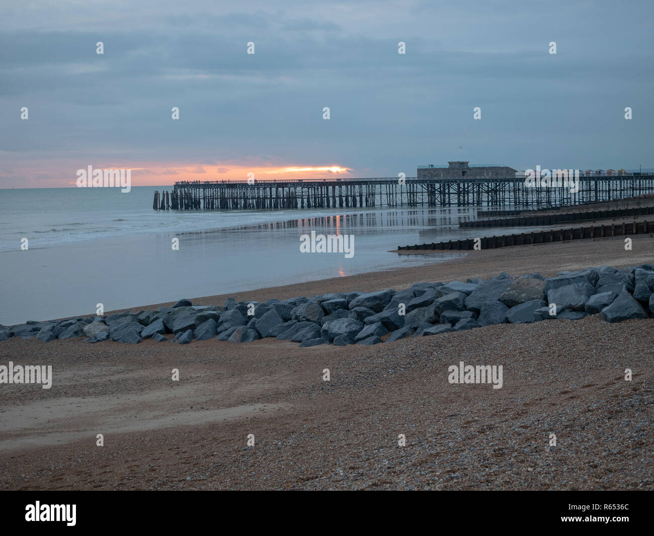 Hastings Pier in inverno freddo La luce al tramonto con la spiaggia, i frangiflutti e barriera di roccia in primo piano Foto Stock