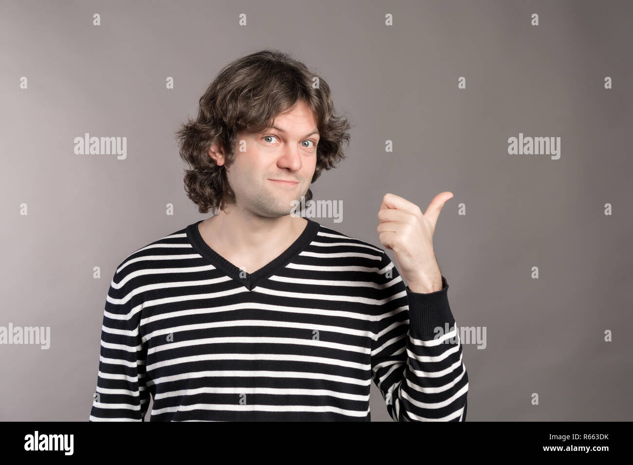 Giovane uomo bello sorridendo felice cercando di fotocamera e rivolta verso il lato con il pollice. Studio shot di allegro capelli scuri guy mostra nuova voce, pone ag Foto Stock