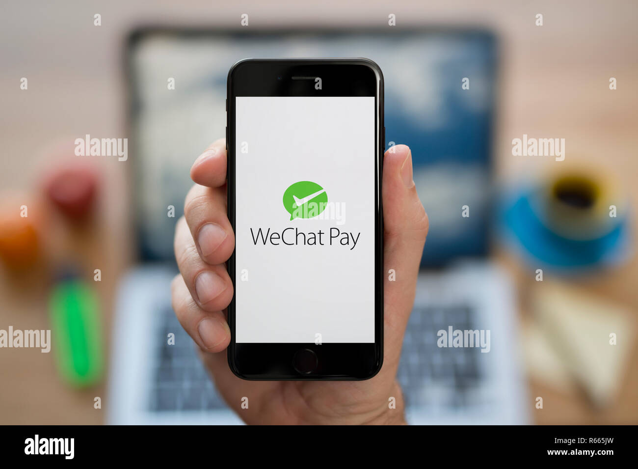 Un uomo guarda al suo iPhone che visualizza il WeChat pagare il logo, mentre se ne sta seduto alla sua scrivania per computer (solo uso editoriale). Foto Stock
