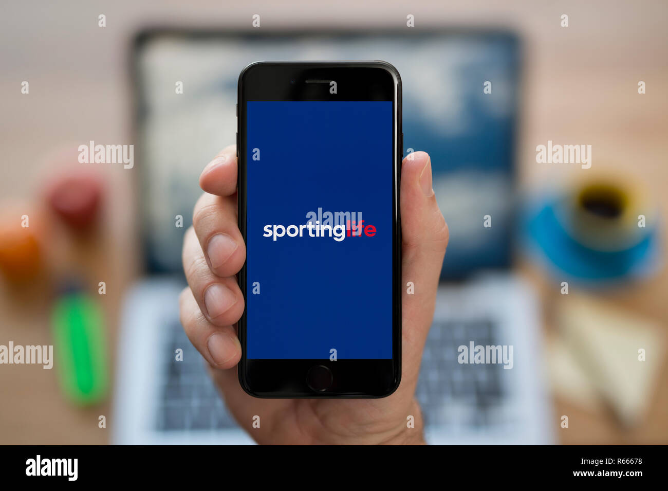 Un uomo guarda al suo iPhone che visualizza lo Sporting Life logo, mentre se ne sta seduto alla sua scrivania per computer (solo uso editoriale). Foto Stock