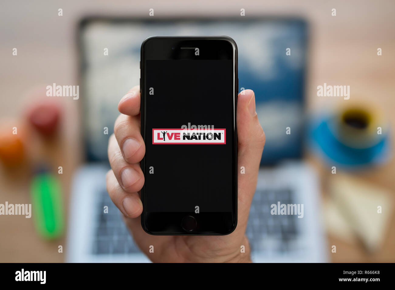 Un uomo guarda al suo iPhone che visualizza il Live Nation logo, mentre se ne sta seduto alla sua scrivania per computer (solo uso editoriale). Foto Stock