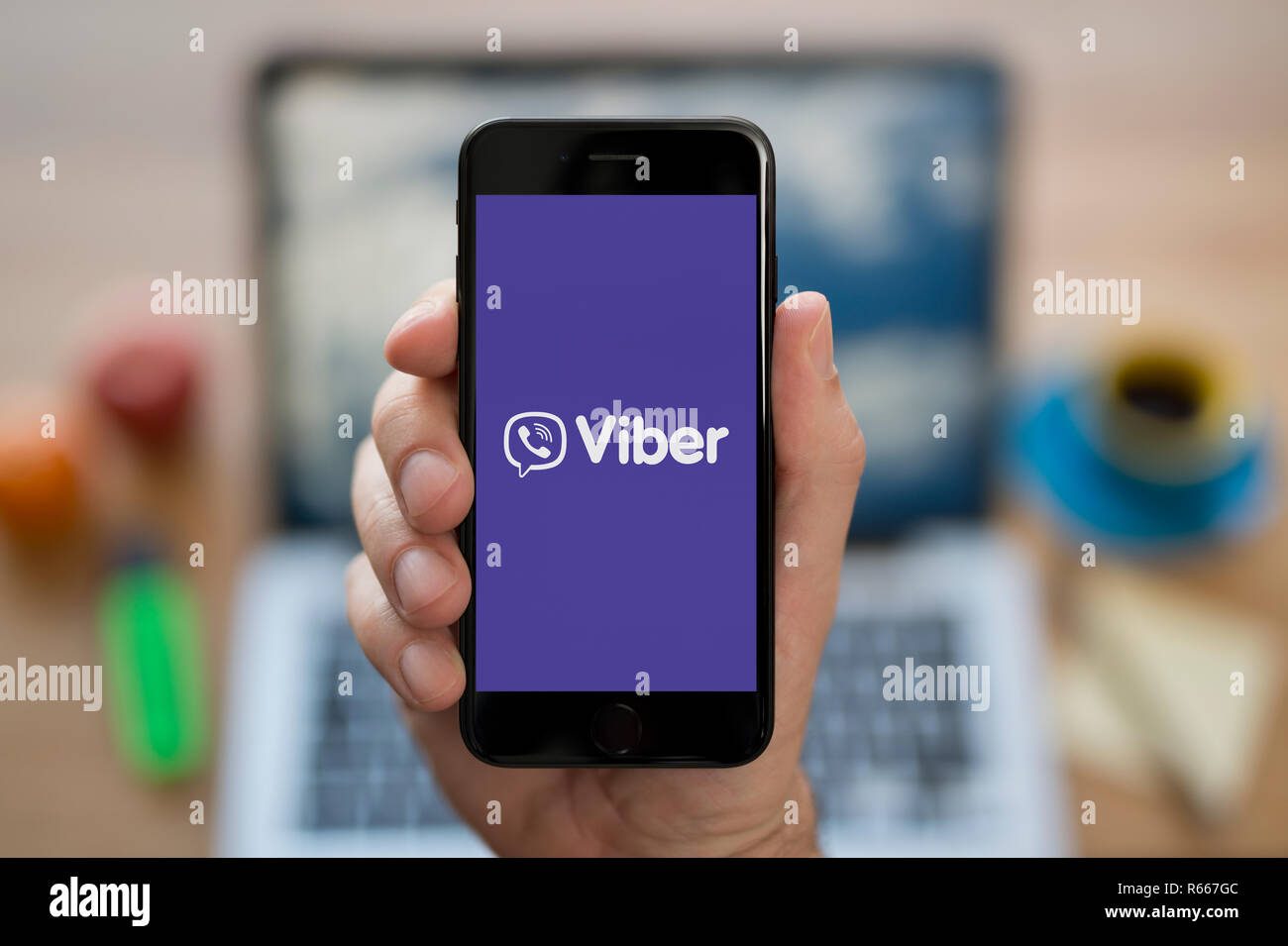 Un uomo guarda al suo iPhone che visualizza il logo Viber, mentre se ne sta seduto alla sua scrivania per computer (solo uso editoriale). Foto Stock