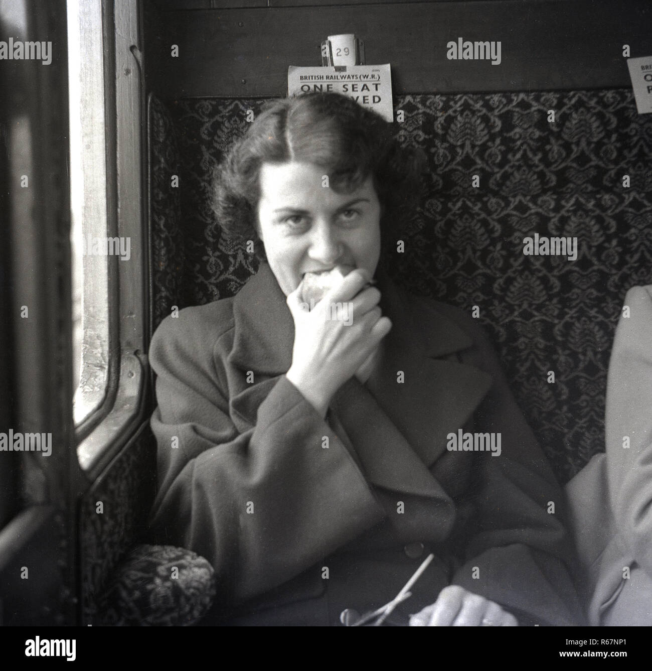 Degli anni Cinquanta, storico, una giovane signora seduta nel suo cappotto in una tradizionale stazione vano accanto alla finestra di mangiare un apple. Foto Stock