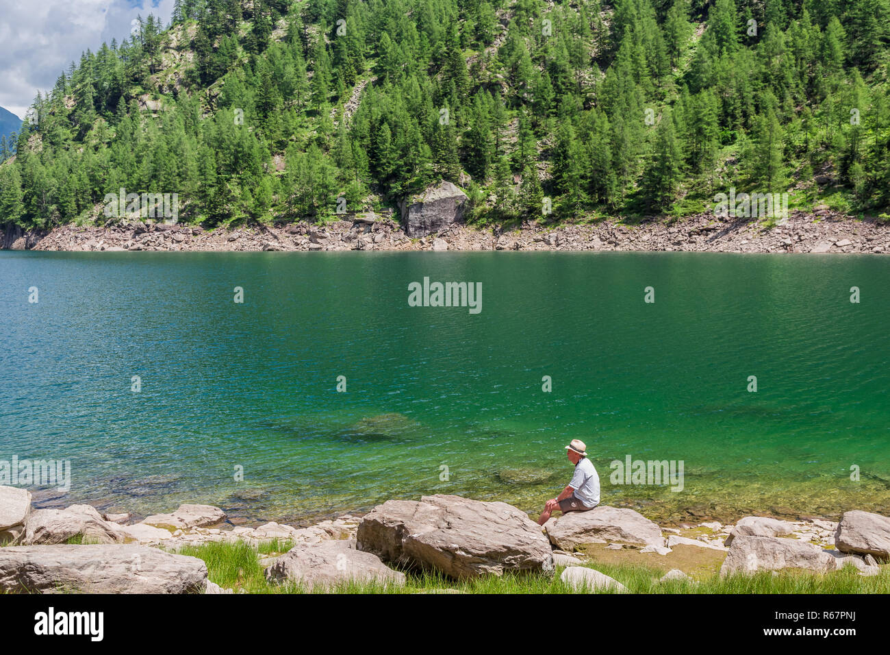 Uomo anziano, indossando un cappello di Panama, seduto da solo su una roccia da un lago. Foto Stock