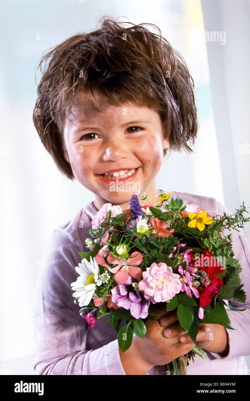Ritratto, Indoor, schmutziges kleines Maedchen mit kurzen braunen Haaren haelt laechelnd einen bunten Blumenstrauss in den Haenden Foto Stock