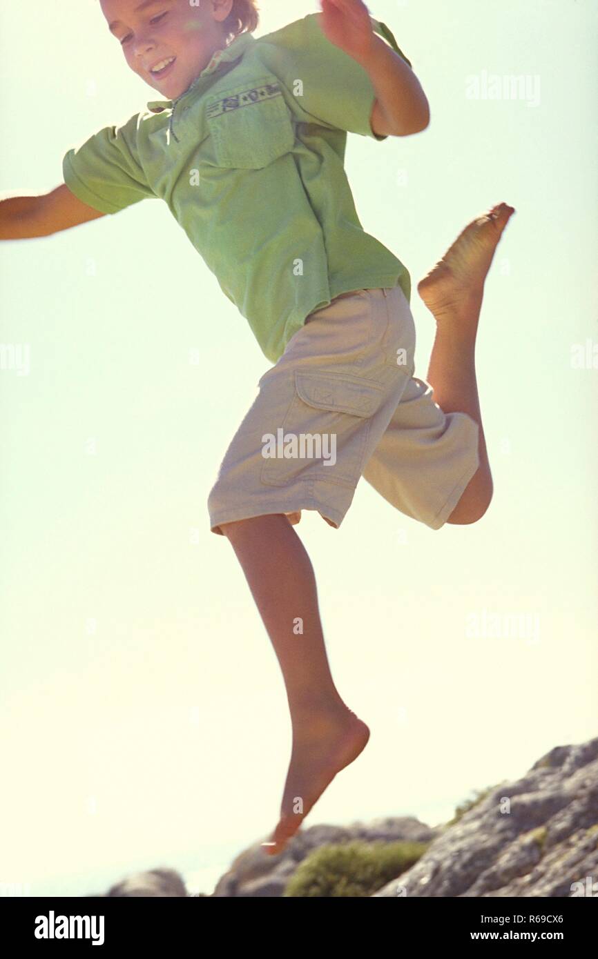 Ganzfigur, 10 Jahre alter blondr Junge, bekleidet mit heller knielanger flessibile, gruenem Hemd springt barfuss ueber Felsen Foto Stock