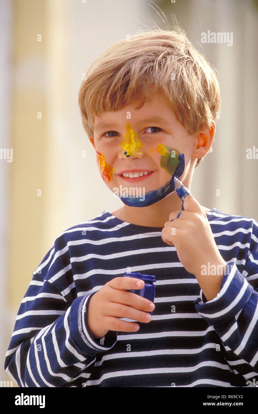 Ritratto, all'aperto, blondr Junge, 6 Jahre alt, bekleidet mit blau-weiss gestreiftem T-Shirt bemalt sich das Gesicht mit Fingerfarben Foto Stock
