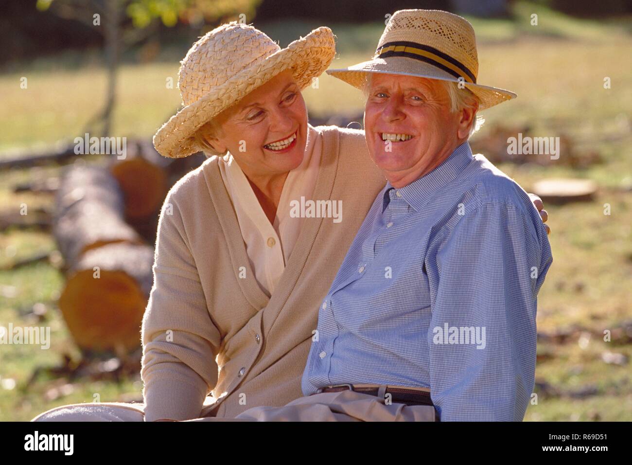 Ritratto, Halbfigur, Seniorenpaar bekleidet mit heller Kleidung und Stohhueten sitzt gluecklich zusammen auf einer Lichtung im Wald Foto Stock