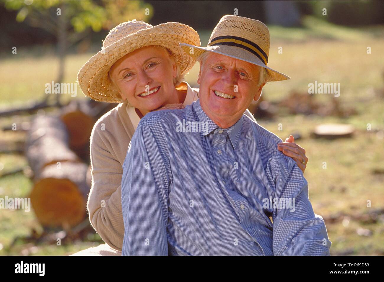 Ritratto, Halbfigur, Seniorenpaar bekleidet mit heller Kleidung und Stohhueten sitzt gluecklich zusammen auf einer Lichtung im Wald Foto Stock