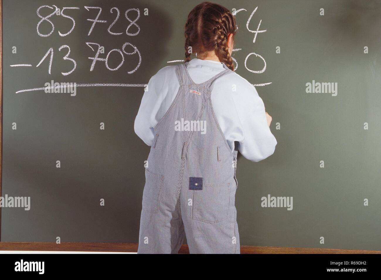 Ritratto, Innenraum, 10-jaehriges Maedchen bekleidet mit Jeanslatzhose steht an der Tafel und rechnet eine Mathematik Aufgabe Foto Stock