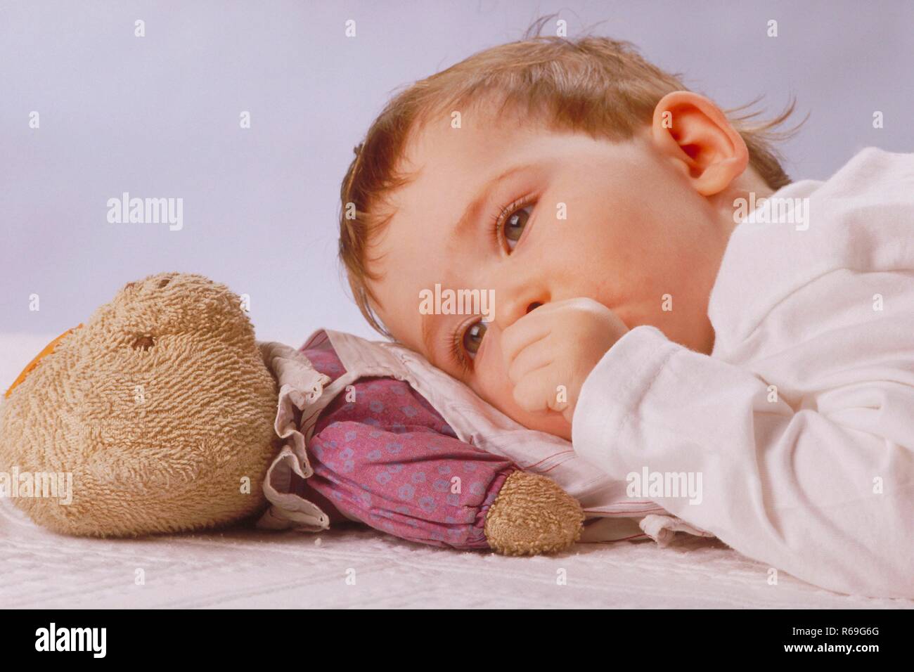 Ritratto, Halbfigur, Innenraum, 2-jaehriges rothaariges Maedchen liegt im Schlafanzug mit dem Daumen im Mund auf ihrem Kuscheltier Foto Stock