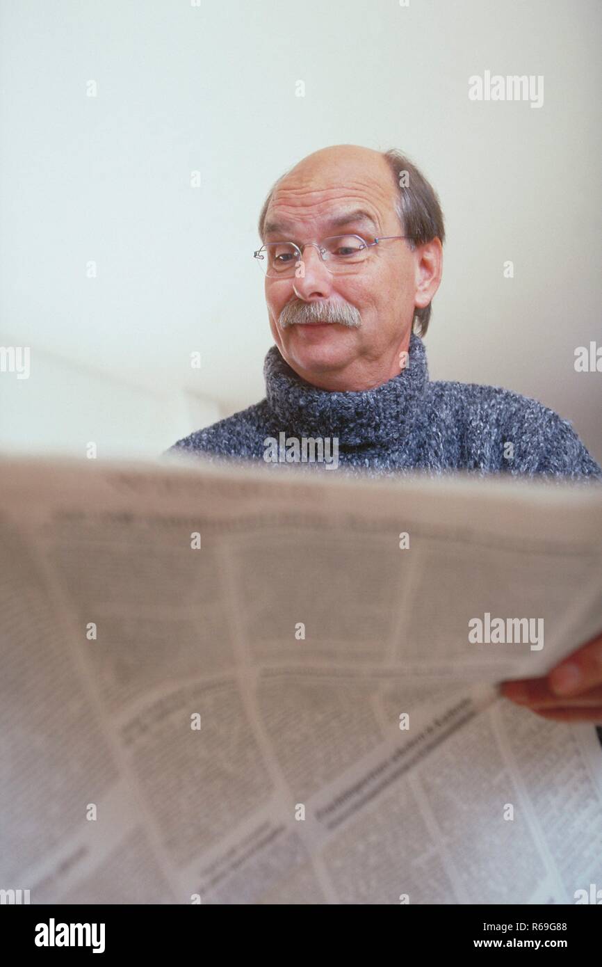 Ritratto, Innenraum, Mann, Mitte 50, mit Halbglatze, Schnauzbart und randloser Brille bekleidet mit grauen dickem Pullover interessiert liest in der Zeitung Foto Stock
