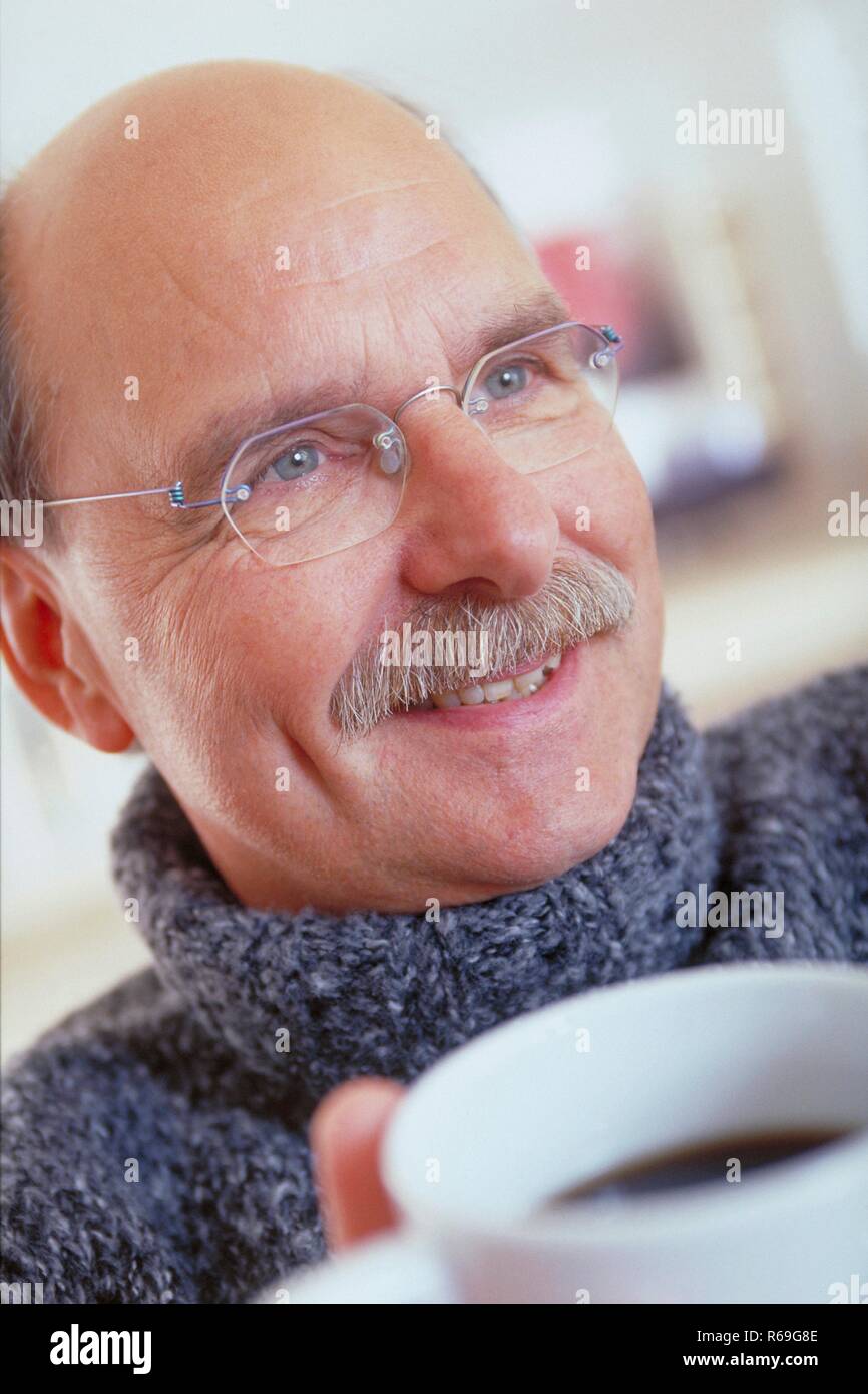 Ritratto, Innenraum, Nahaufnahme, Mann, Mitte 50, mit Halbglatze, Schnauzbart und randloser Brille bekleidet mit grauen dickem Pullover freut sich ueber eine Tasse Kaffee Foto Stock