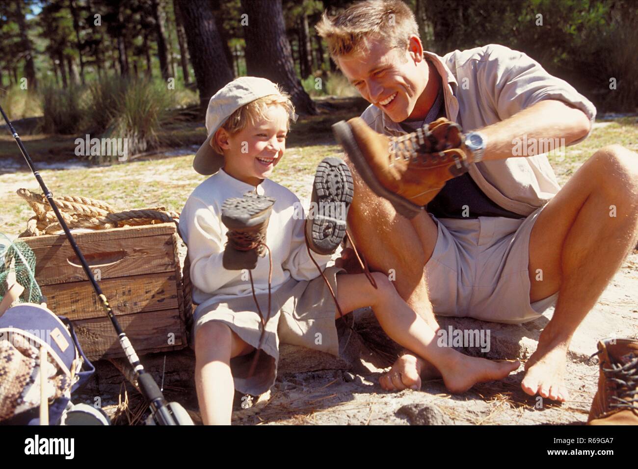 All'aperto, 6 Jahre alter blondr Junge sitzt mit seinem Vater barfuss auf Steinen im Wald und halten gluecklich ihre Wanderschuhe in den Haenden Foto Stock