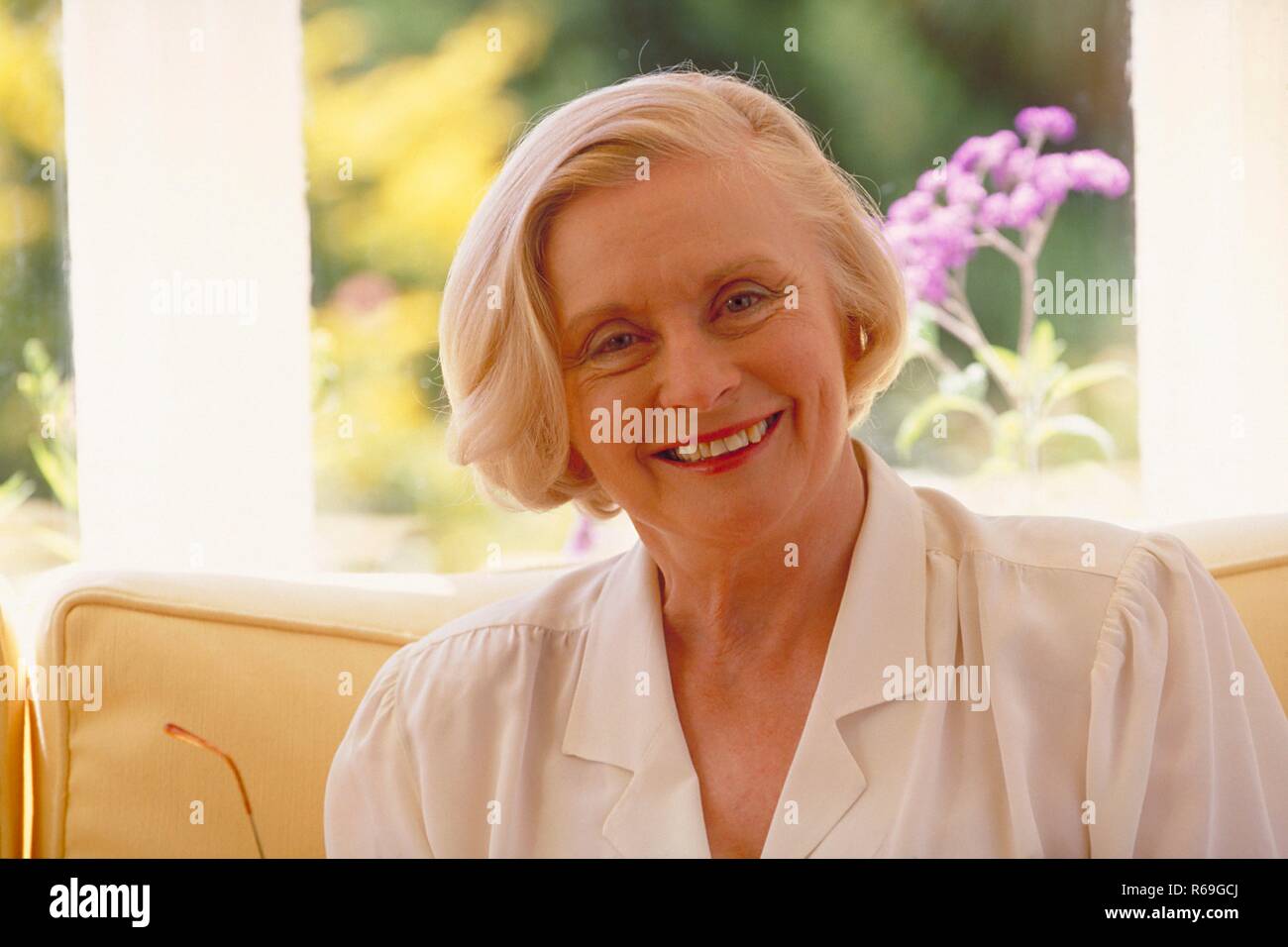 Interni, ritratti, blonde Frau Mitte 60 bekleidet mit weisser Bluse sitzt im Wohnzimmer vor dem Fester mit Blick in den Garten auf dem divano Foto Stock