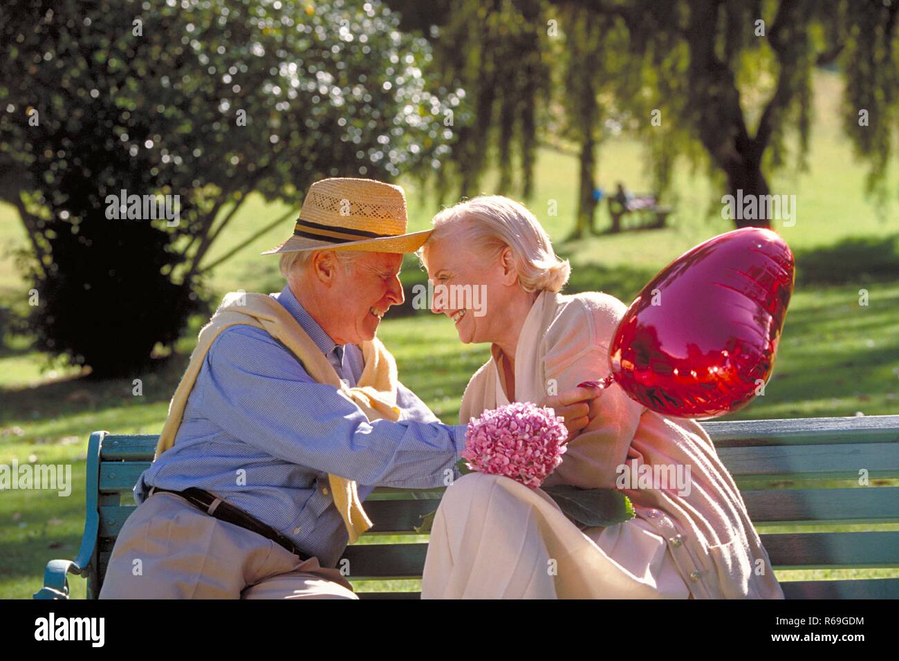 Parkszene, Outdoor, Halbfigur, Seniorenpaar bekleidet mit heller Kleidung, er mit Strohhut, sitzt flirtend Mit einem roten herzfoermigen Luftballon und einem Fliederzweig auf einer Parkbank Foto Stock