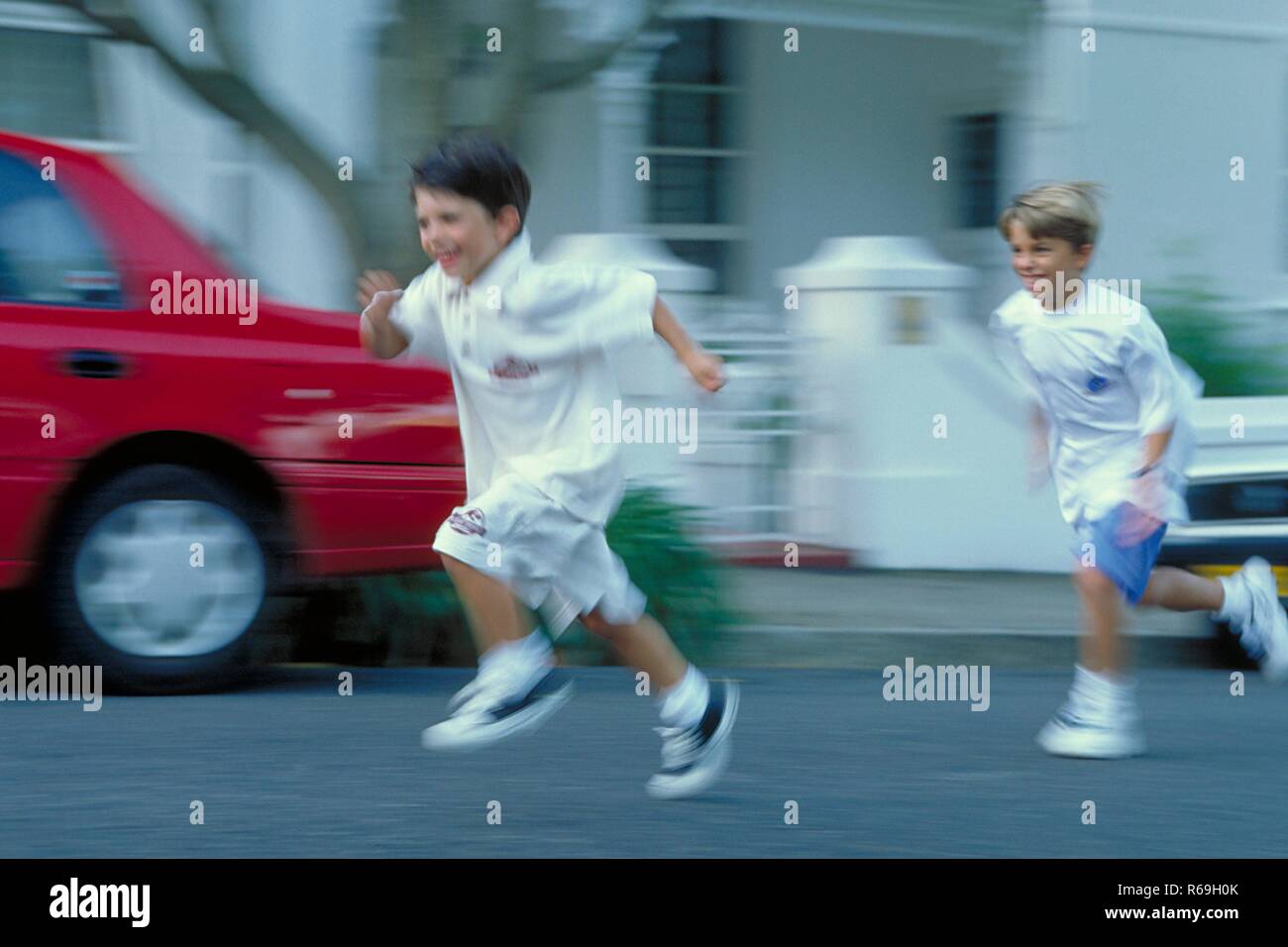 Ritratto, Ganzfigur, zwei 7-8 Jahre alte Jungen bekleidet mit weissen T-Shirts und Shorts laufen un einem roten Auto vorbei die Strasse entlang Foto Stock