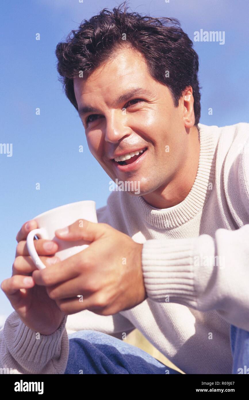 Strandszene, Ritratto, junger Mann mit schwarzen Haaren und braunen Augen, bekleidet Mit einem hellem Pullover und Jeans, sitzt entspannt mit einer weissen Tasse Kaffee in der mano am Strand Foto Stock