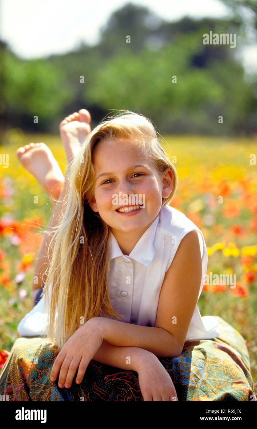 Ritratto, laechelndes Maedchen mit langen Haaren blondn, 12 Jahre, mit weisser Bluse aermellosen sitzt auf einer mit Klatschmohn und gelben Blueten uebersaeten Wiese Foto Stock