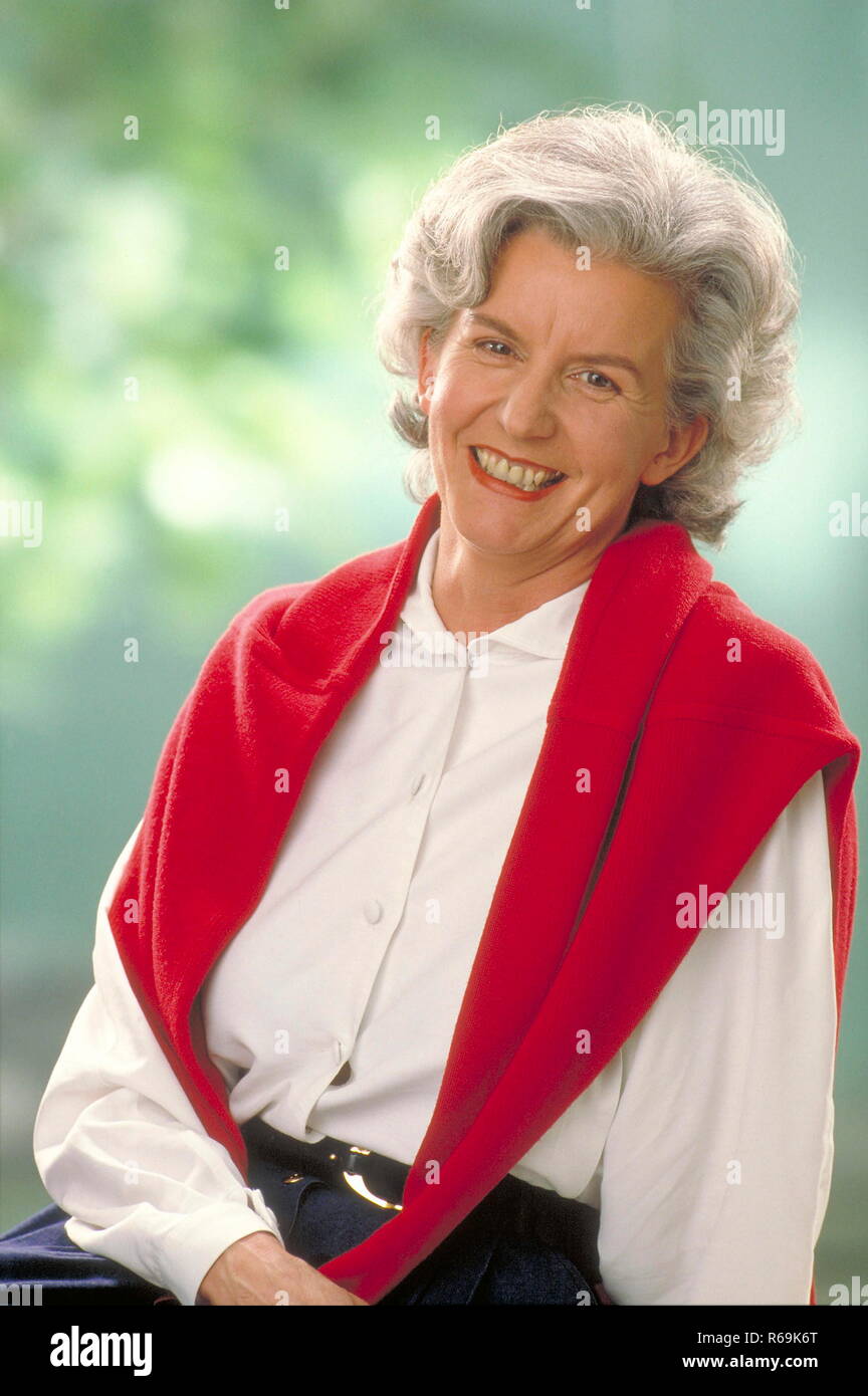 Ritratto, lachende Frau mit grauem Haar im Gruenen, ca. 60 Jahre, traegt weisse Bluse und einen roten Pullover ueber der Schulter Foto Stock