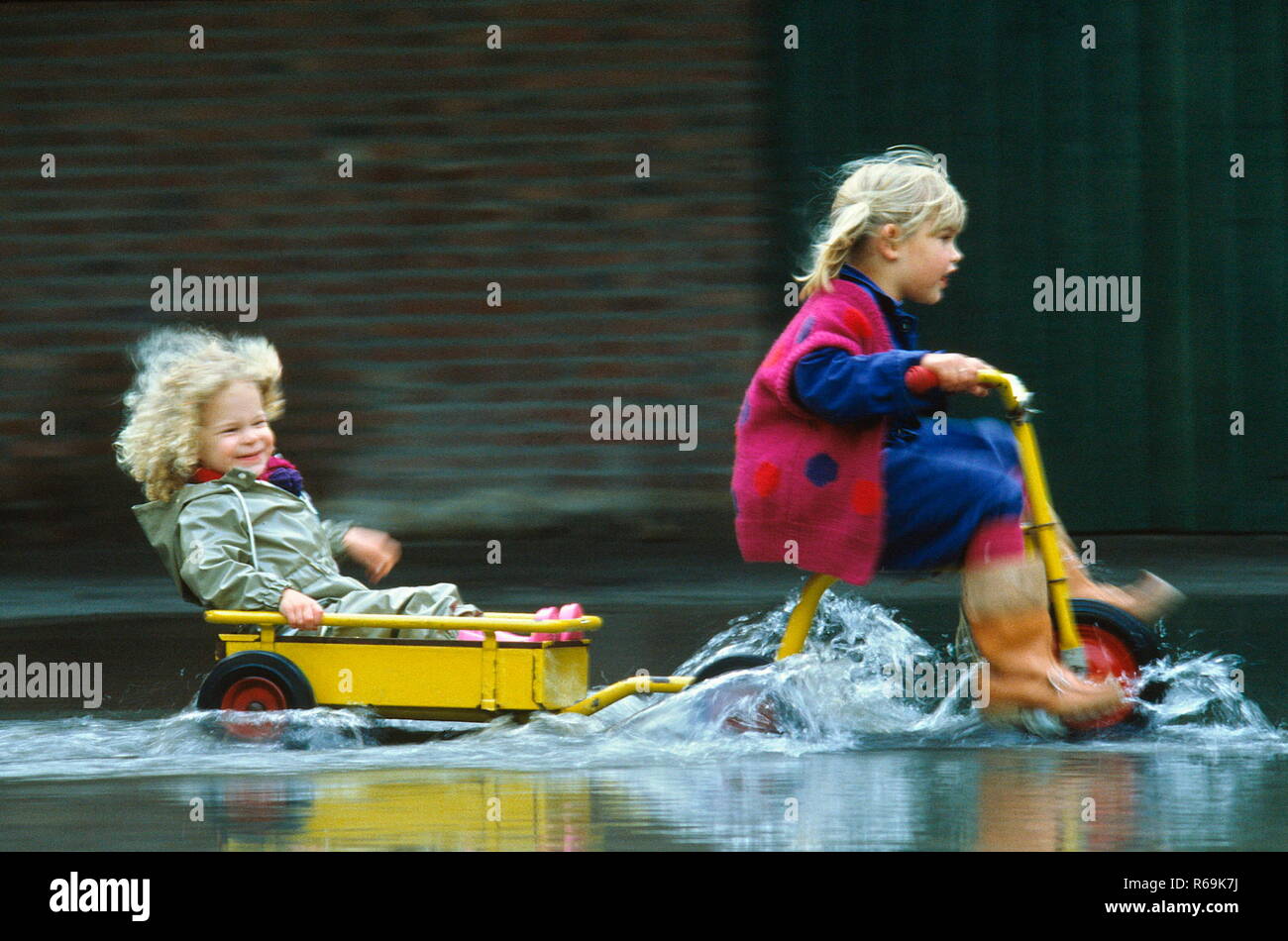 Ritratto, Herbstszene, bionde Maedchen, 4 Jahre, zieht mit gelbem Dreirad ihre in einem Anhaenger sitzende kleine Schwester, 2 Jahre, durch eine tiefe Pfuetze Foto Stock