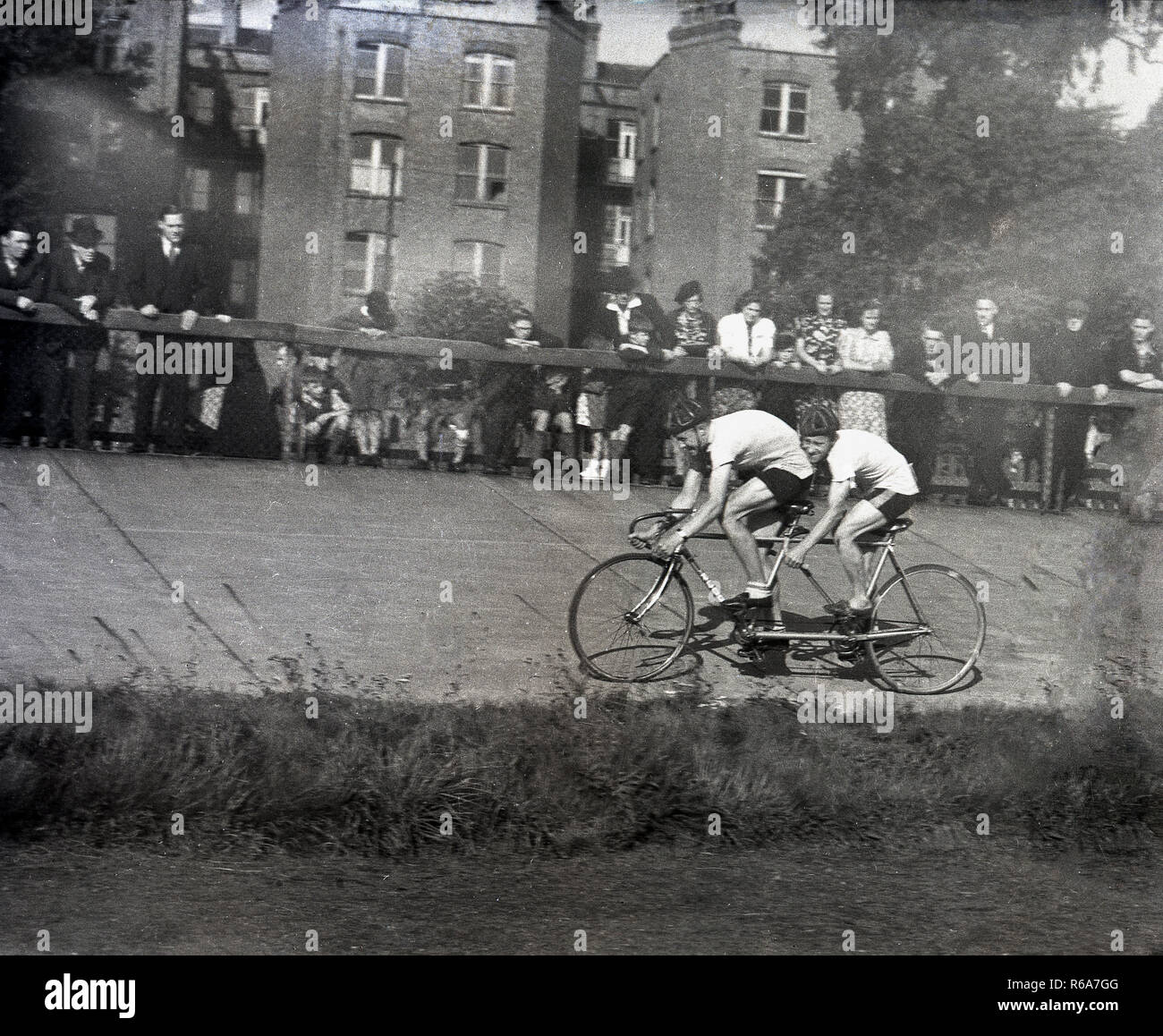 1940s, storico, due corridori ciclisti su un tandem bike fuori in una gara ciclistica su un calcestruzzo rialzata in via, guardato dagli spettatori in piedi dietro un recinto, Inghilterra, Regno Unito. Foto Stock