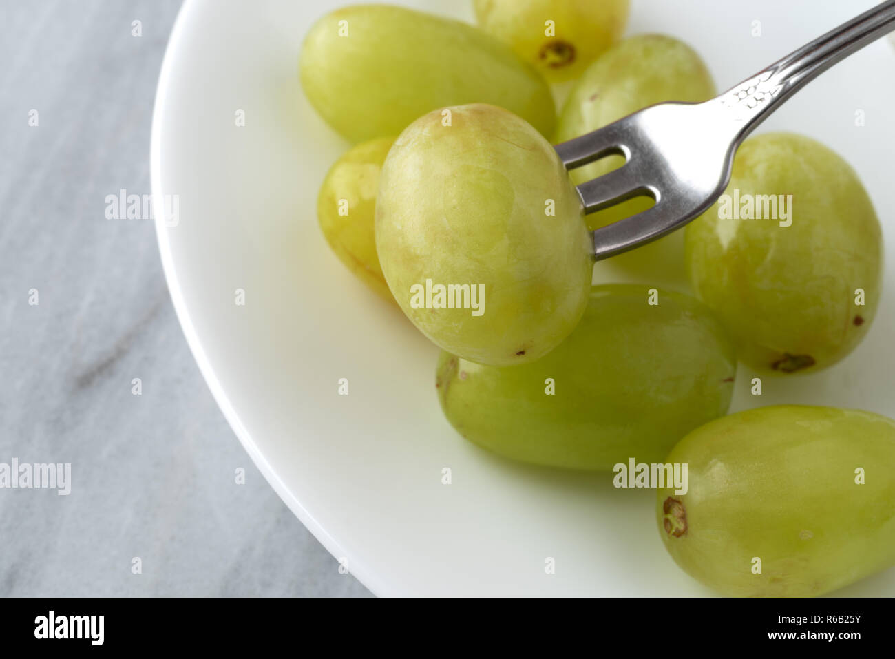 Chiudere la vista di un serving di uva verde con una forcella skewering un pezzo di frutta su una piastra bianca in cima a un banco di marmo alto. Foto Stock