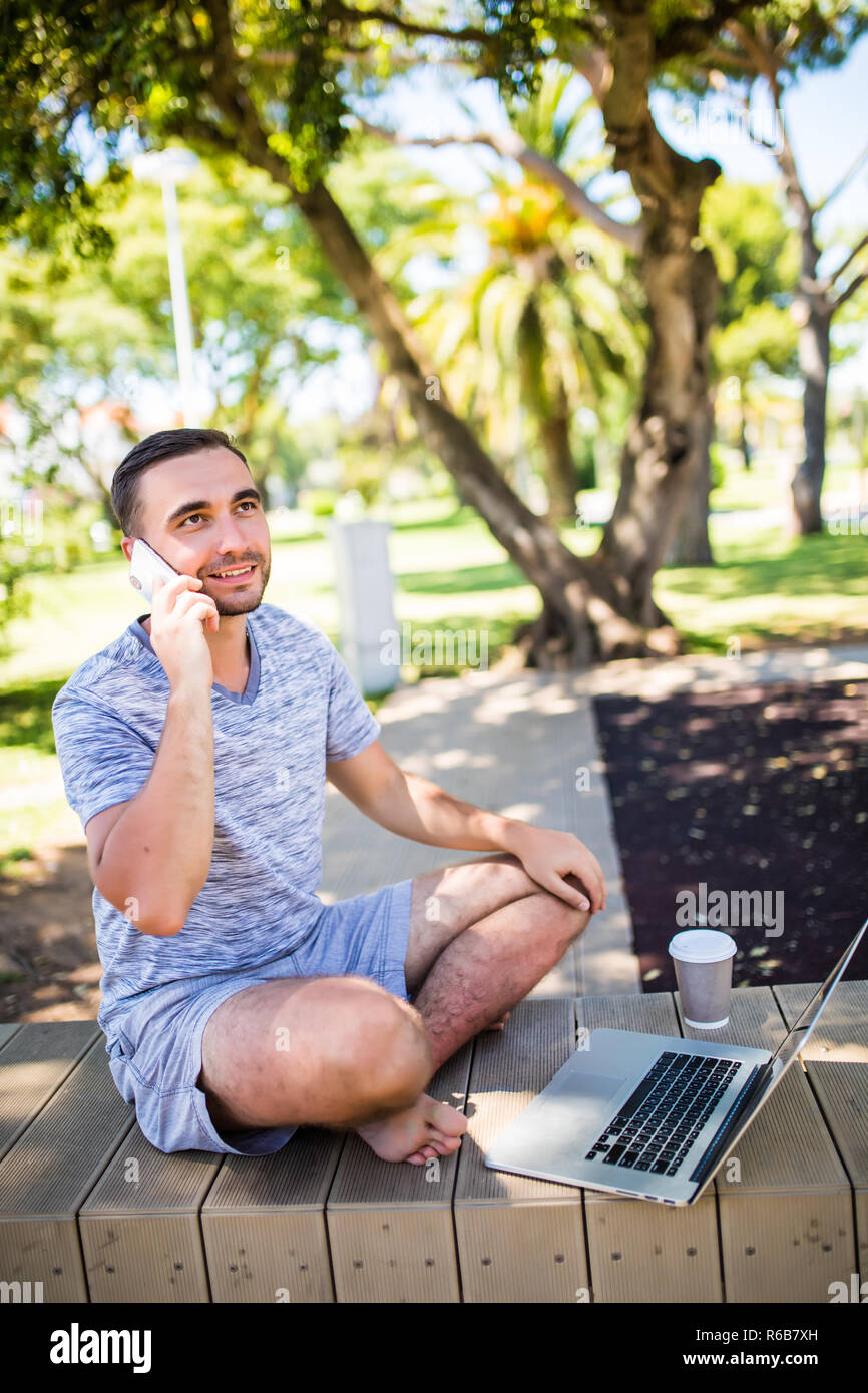 Copywriter sorridente parlando al telefono cellulare durante il lavoro a distanza sul computer portatile, seduti nel parco della città nel giorno d'estate. Hipster guy chiama tramite cellph Foto Stock