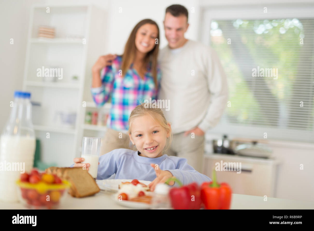 Bellissima bambina avente la colazione nella cucina domestica. La sua felicità ai genitori di stare dietro a. Guardando alla fotocamera. Foto Stock