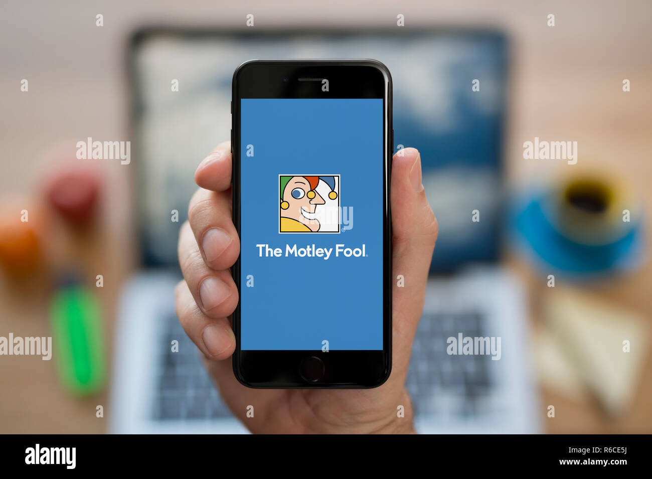 Un uomo guarda al suo iPhone che visualizza il Motley Fool logo, mentre se ne sta seduto alla sua scrivania per computer (solo uso editoriale). Foto Stock