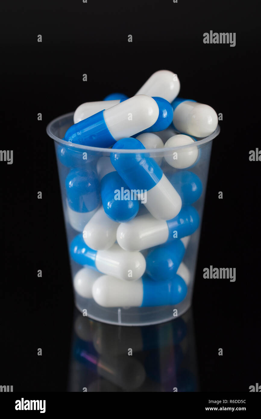 Big pharma farmaco concetto. Blu e bianco capsule pillole nel bicchiere graduato in plastica / coppa di erogazione sulla superficie riflettente. Possibile metafora di NHS. Foto Stock