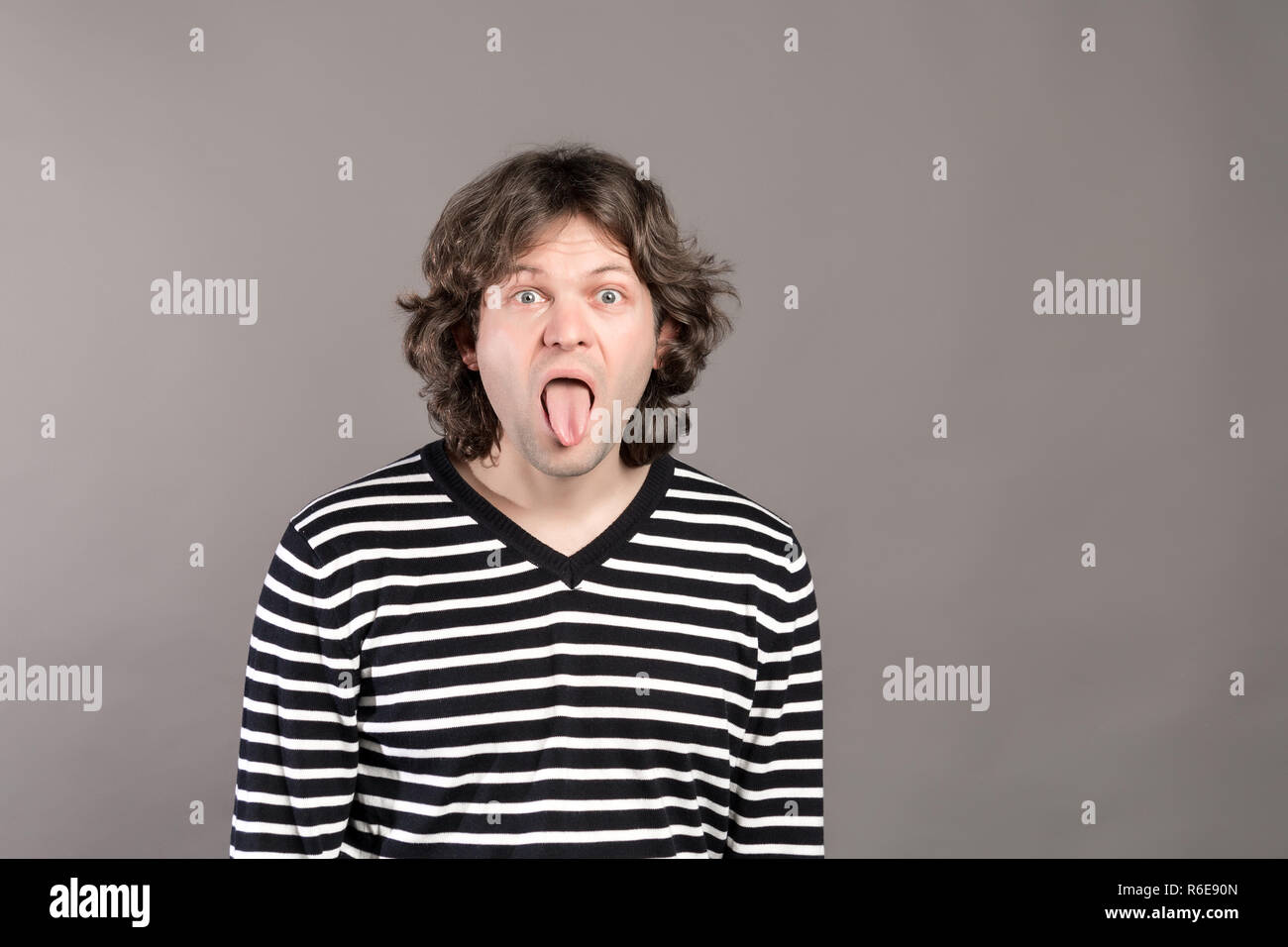 Ritratto di funny shaggy pazzo uomo bello in maglione a strisce guardando la fotocamera con grande bocca aperta e la lingua di fuori, teases. Ampia aprì gli occhi indoor studio shot, isolata su uno sfondo grigio. Foto Stock