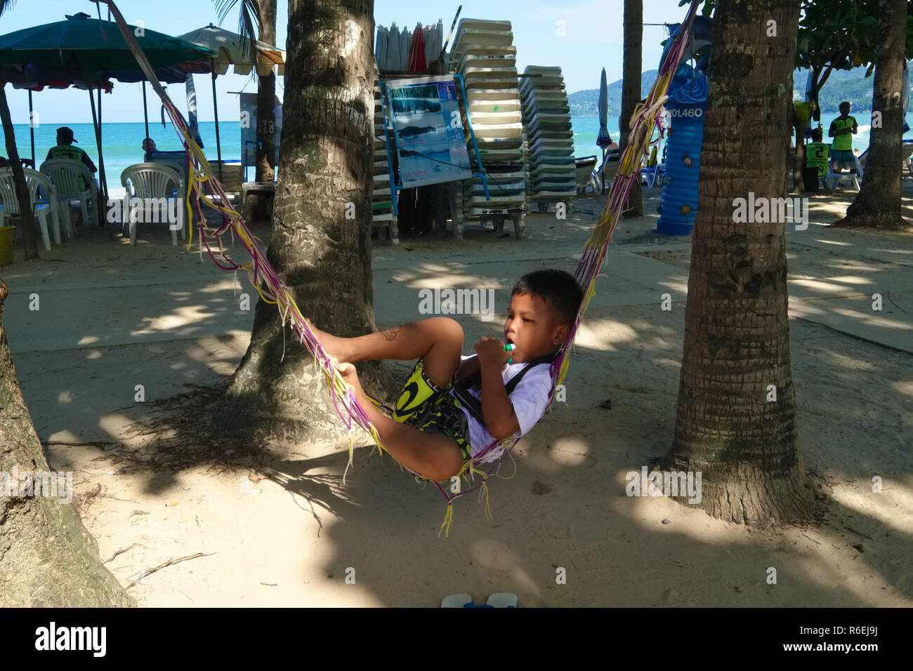 Un ragazzo locale a Patong Beach, Phuket, Thailandia, prende vita facile in una amaca, mentre i suoi genitori eseguire un business sulla spiaggia Foto Stock