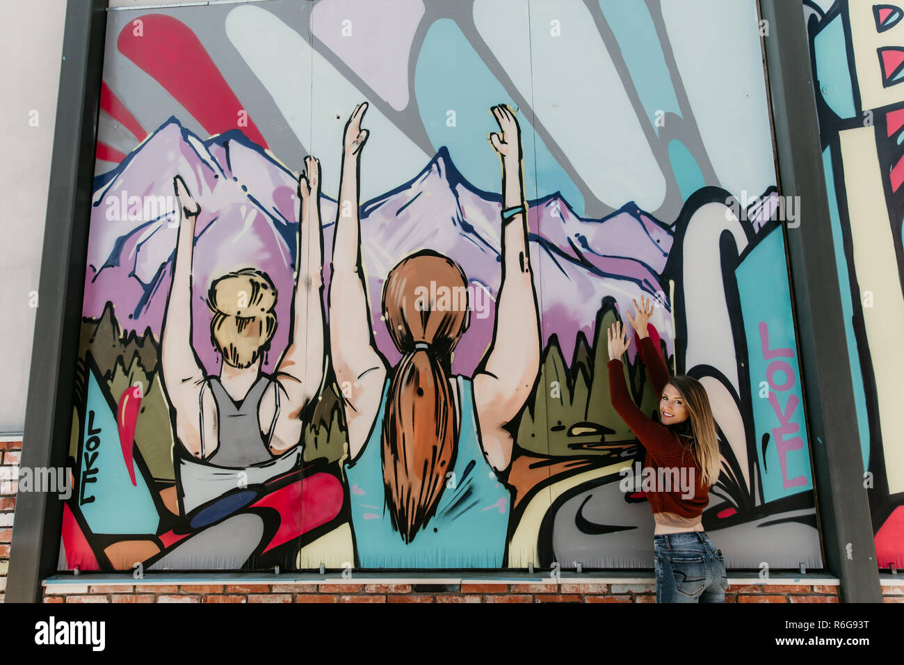 Piuttosto carino bionda e giovane donna adulta al di fuori nella stagione fredda Downtown Urban Street Art District seduti di fronte vibrante coloratissimo graffito Art Wall Foto Stock