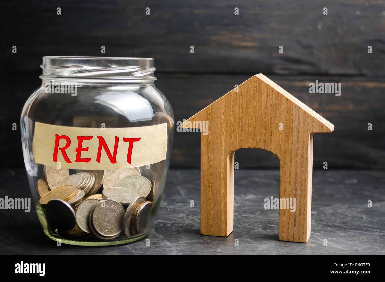 Vaso di vetro con le monete e la scritta "nt' e una piccola casa in legno. Affitto di immobili, appartamenti. i servizi di un agente immobiliare. abitazioni a prezzi accessibili, r Foto Stock