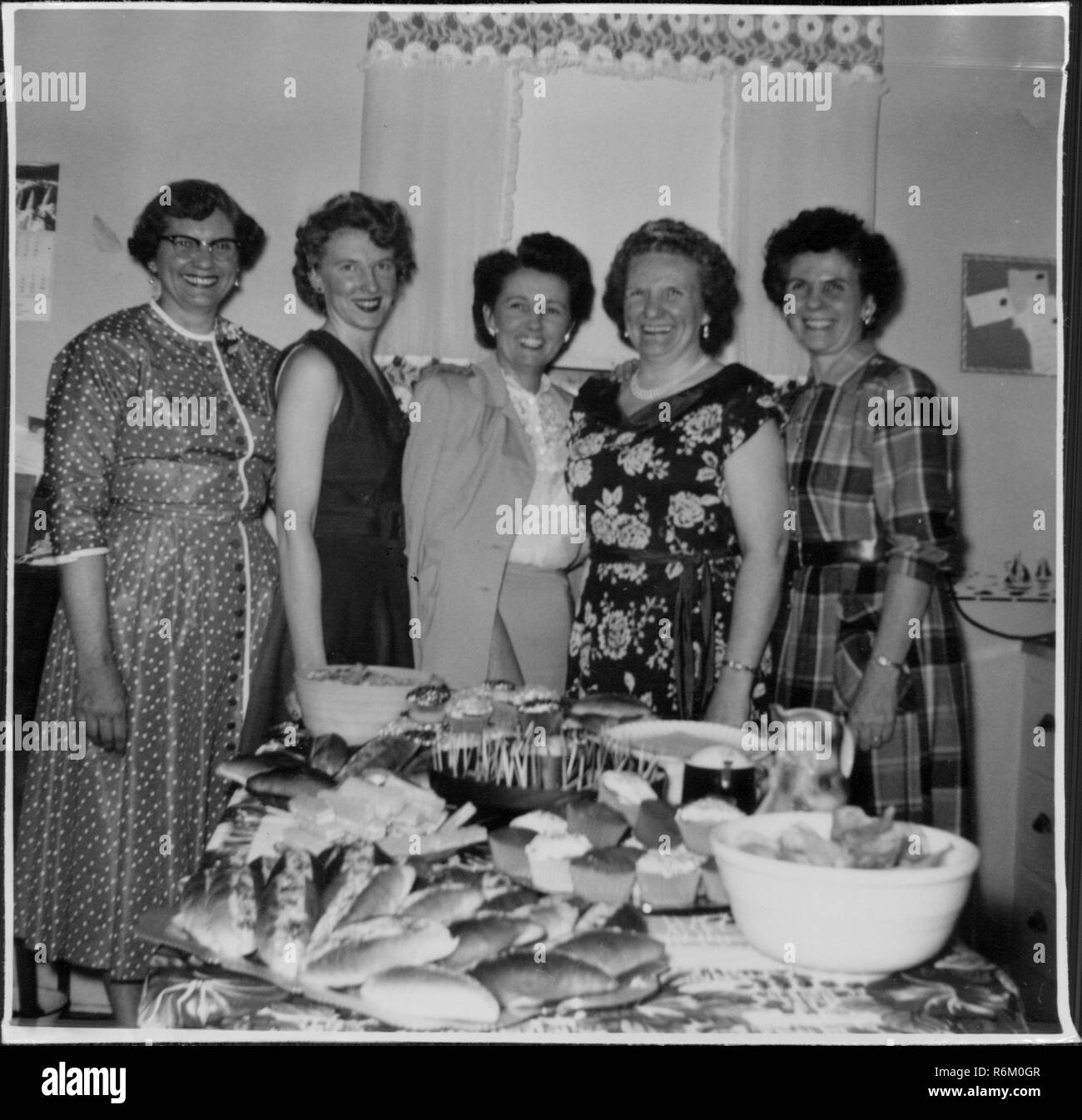 Un gruppo delle casalinghe di età variabili, indossando abiti, in piedi in una casa suburbana e in posa per una fotografia con un tavolo ricoperto nella tradizionale festa alimenti, incluso scheggiature, panini e casseruole dip, 21 aprile 1954. () Foto Stock